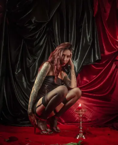 Moody portrait of a tattooed woman in a black dress sitting against red and black satin drapes by candlelight.