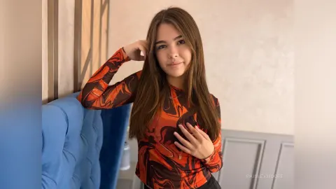 PaisleyMoore — PaisleyMoore with long brown hair poses in an orange and black patterned long-sleeve top while leaning against a blue headboard.