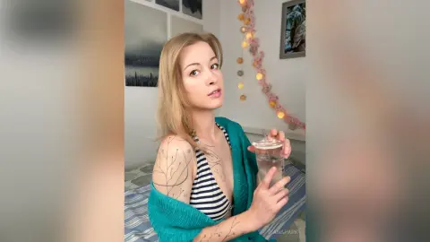 Blonde woman in a striped bikini and teal cover-up holding a drink in a bright, cozy indoor setting.