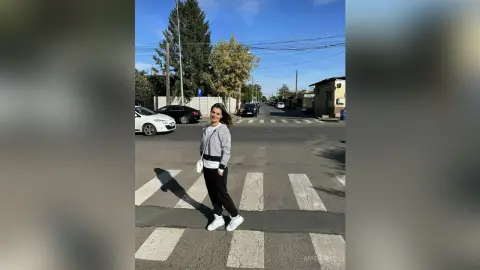Smiling woman in a grey sweater and black trousers standing on a city street crosswalk on a sunny day.