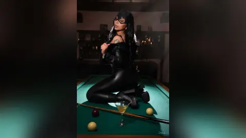 Woman in black leather catsuit and mask sitting on a pool table in a moody, dimly lit lounge setting.