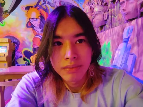 Young man in a white t-shirt posing in front of colorful anime mural art in a vibrant, neon-lit room.