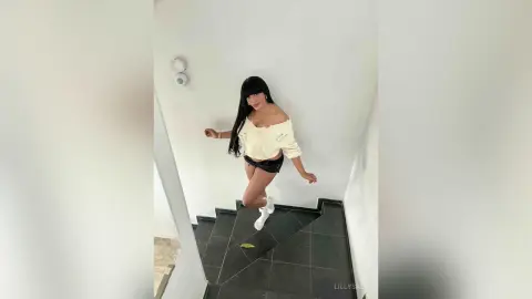 Young woman in a white off-the-shoulder top and black shorts posing in a minimalist white staircase setting.