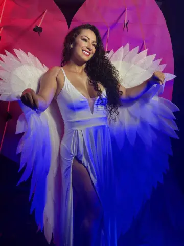 Smiling woman in a white flowing dress and large angel wings posing against a pink and blue studio background.