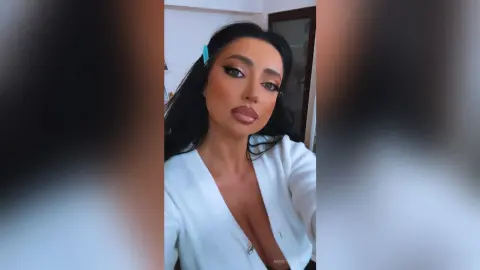 Glamorous woman with dark hair and heavy makeup wearing a white V-neck blouse in a bright indoor setting.