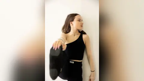 Young woman in a black tank top and leggings posing against a white wall in a minimalist indoor setting.