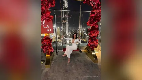 Elegant woman in a white gown sitting on a swing decorated with red flowers overlooking a city skyline at night.