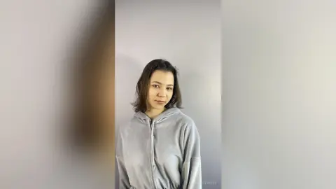 Young woman in a cozy grey zip-up hoodie posing against a neutral studio background with a calm expression.