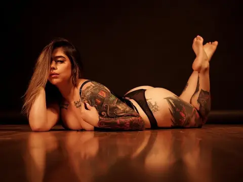 Nomohah_sainz lies on her stomach on a dark floor, showing off her tattoos and black lingerie while looking at the camera.