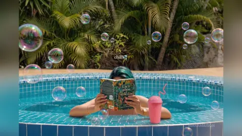 Relaxed woman in a blue bikini reading a book in a tropical swimming pool surrounded by floating bubbles.