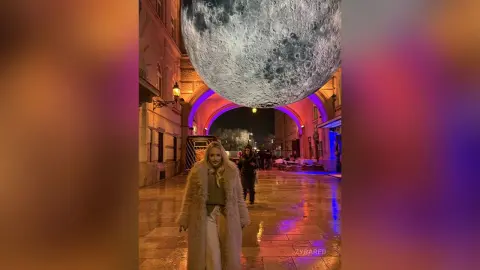 Blonde woman in a fuzzy beige coat walking through a neon-lit cobblestone alley under a giant moon.