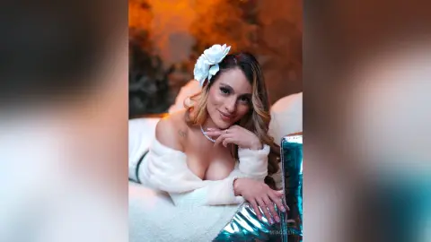 Smiling woman in a white fuzzy sweater and flower hair accessory relaxing in a cozy, warm setting.