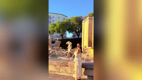 Stylish woman in a white summer dress standing by a classical stone fountain in a sunny European plaza.