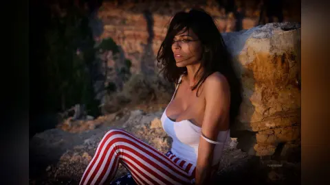 Beautiful woman in white top and red striped patriotic pants sitting against a rock in a canyon at sunset.