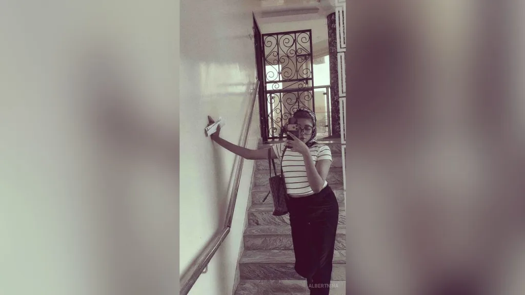 AlbertNina — AlbertNina stands in a hallway wearing a striped shirt and black skirt while holding a phone for a selfie.