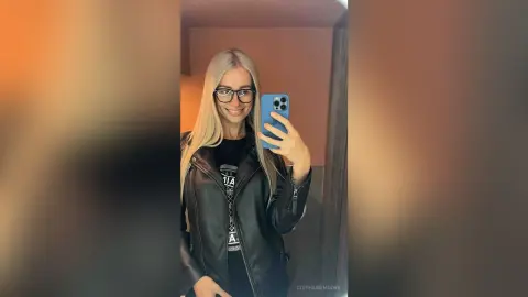 Smiling blonde woman in black leather jacket and glasses taking a mirror selfie in a modern room.
