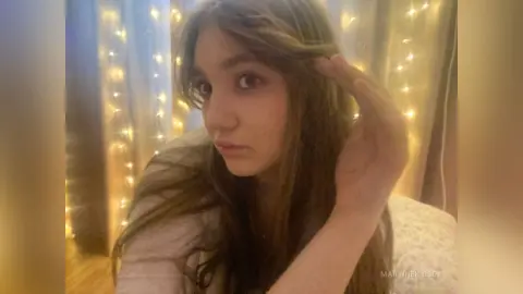MartineKibbey poses with her long brown hair, looking at the camera against a background of warm string lights.