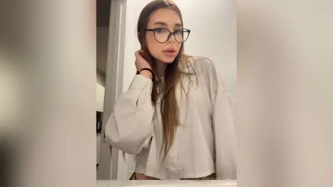 Young woman with long hair and glasses wearing a white oversized button-down shirt in a bright indoor setting.