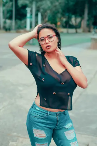 NiaMorelli poses outdoors in a sheer black top and ripped blue jeans, running her hand through her dark hair while wearing red glasses.