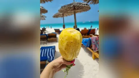 Hand holding a fresh pineapple slice at a sunny tropical beach resort with straw umbrellas.