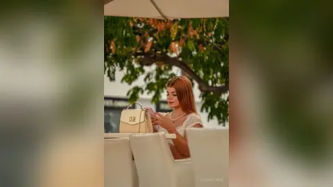 Redheaded woman in a white blouse using her phone at an outdoor cafe with a beige handbag on the table.
