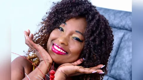 Smiling Black woman with curly hair and red lipstick posing happily in a cozy indoor setting.