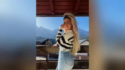 Anita poses outdoors in a striped long-sleeve top and light blue denim jeans against a mountain backdrop.