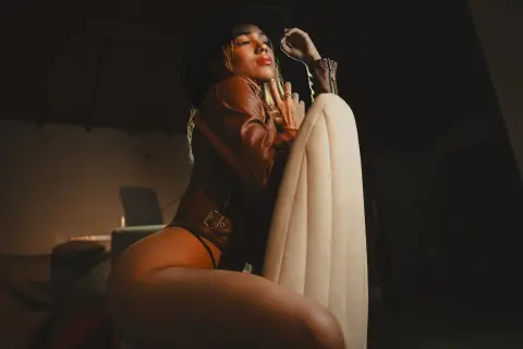 Stylish woman in a brown leather jacket and black hat posing moody in a dimly lit room.