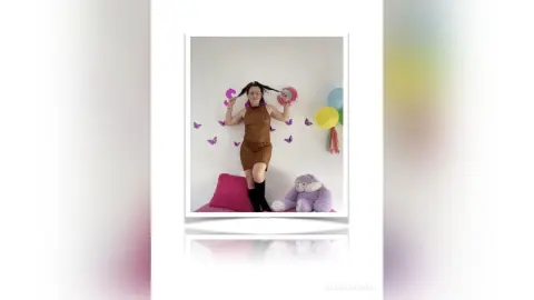 Cheerful woman in a brown sleeveless dress playing with her hair near colorful balloons and a plush toy.