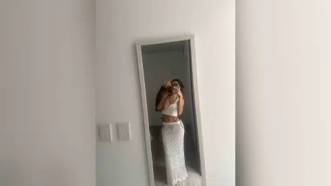 Young woman in a white crop top and long patterned skirt taking a mirror selfie in a minimalist room.