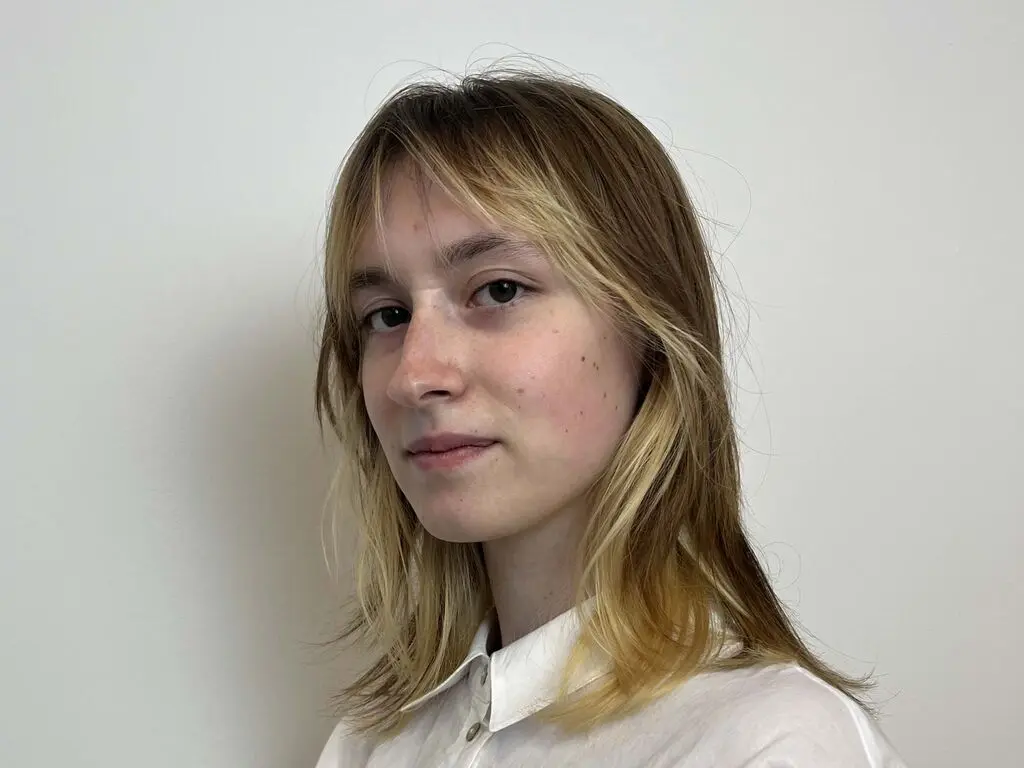 IdaZeccardi looks at the camera with long blonde hair and a white button-down shirt against a plain white background.