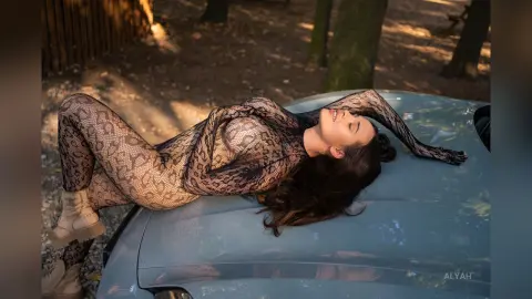 Woman in a snakeskin print bodysuit lounging on a blue car roof in a sunlit forest setting.