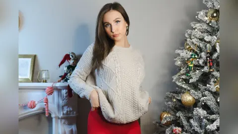 Young woman in a cozy cream cable-knit sweater posing by a decorated Christmas tree in a festive home setting.