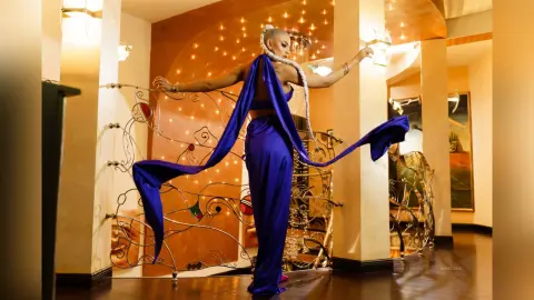 Anastasia stands in a blue silk outfit with long flowing sashes, posing against a decorative metal gate in a warm, lit room.