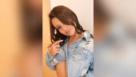 Young woman with long wavy hair wearing a light blue denim jacket in a bright indoor setting.