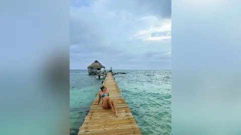 Relaxed woman in a blue bikini lounging on a wooden pier over turquoise tropical ocean water under a cloudy sky.