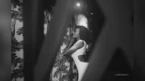 Elegant woman in a white sleeveless dress standing against floral wallpaper in a moody black and white portrait.