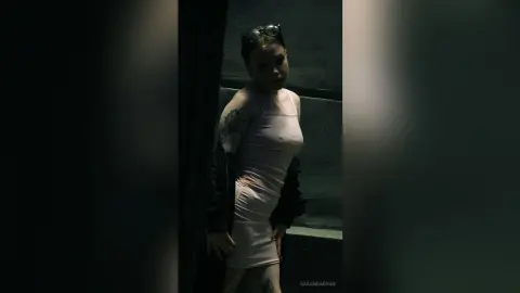 Young woman in a tight white tank top and sunglasses posing in a moody, dark urban setting.