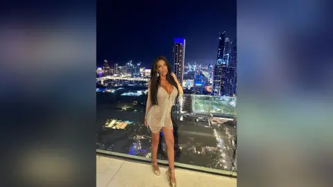 Glamorous woman in a sparkling silver mini dress posing on a luxury rooftop balcony overlooking a night city skyline.