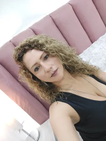 Woman with curly blonde hair wearing a black tank top in a cozy bedroom setting.