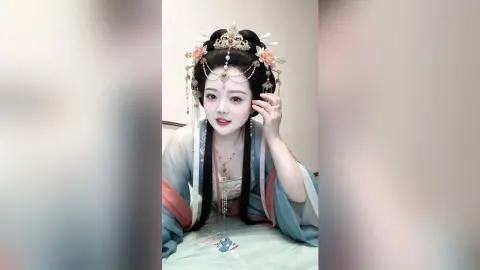 EnyaLin wears an elaborate traditional headpiece and light blue silk robes while leaning forward over a bed.