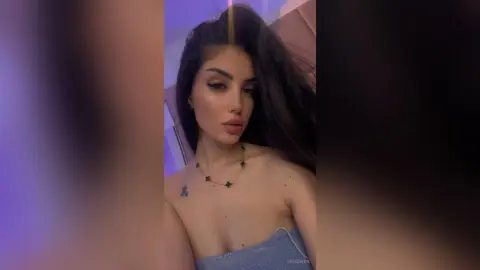 Young woman with long dark hair wearing a silver glitter top and necklace in soft purple lighting.
