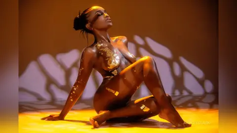 Glamorous woman with gold body glitter sitting in warm cinematic lighting for a luxury beauty concept.