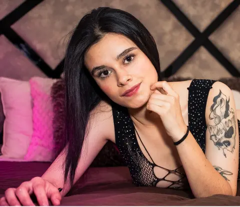 Smiling young woman with long black hair in a black sequined mesh top relaxing on a bed with pink pillows.
