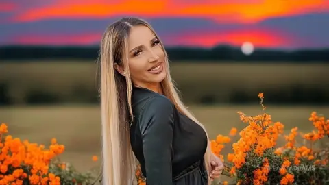 TiffanyAddams smiles while posing outdoors in a field of orange flowers, wearing a black long-sleeved top with her long blonde hair down.