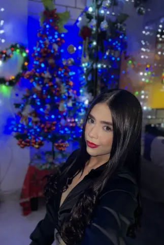 Nomohah_sainz poses in a black shirt with long dark hair, standing in front of a lit blue and red Christmas tree.