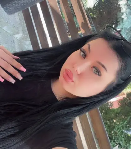 Young woman with long black hair and pink nails wearing a black top in a sunny outdoor garden setting.