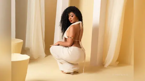 Elegant woman in white backless jumpsuit posing in a minimalist cream studio setting.