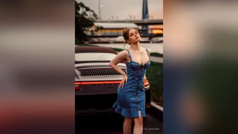 StelaBrown stands next to a dark car wearing a blue denim mini dress with thin straps and a zipper front.