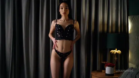 Elegant woman in black floral lace lingerie standing against dark curtains in a moody bedroom setting.
